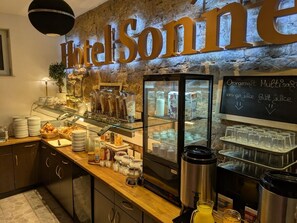A hotel breakfast area with a stone wall, a large sign reading 'Hotel Sonine,' a glass display case with food items, and a chalkboard menu. - Hotel Sonne (Karlsruhe)