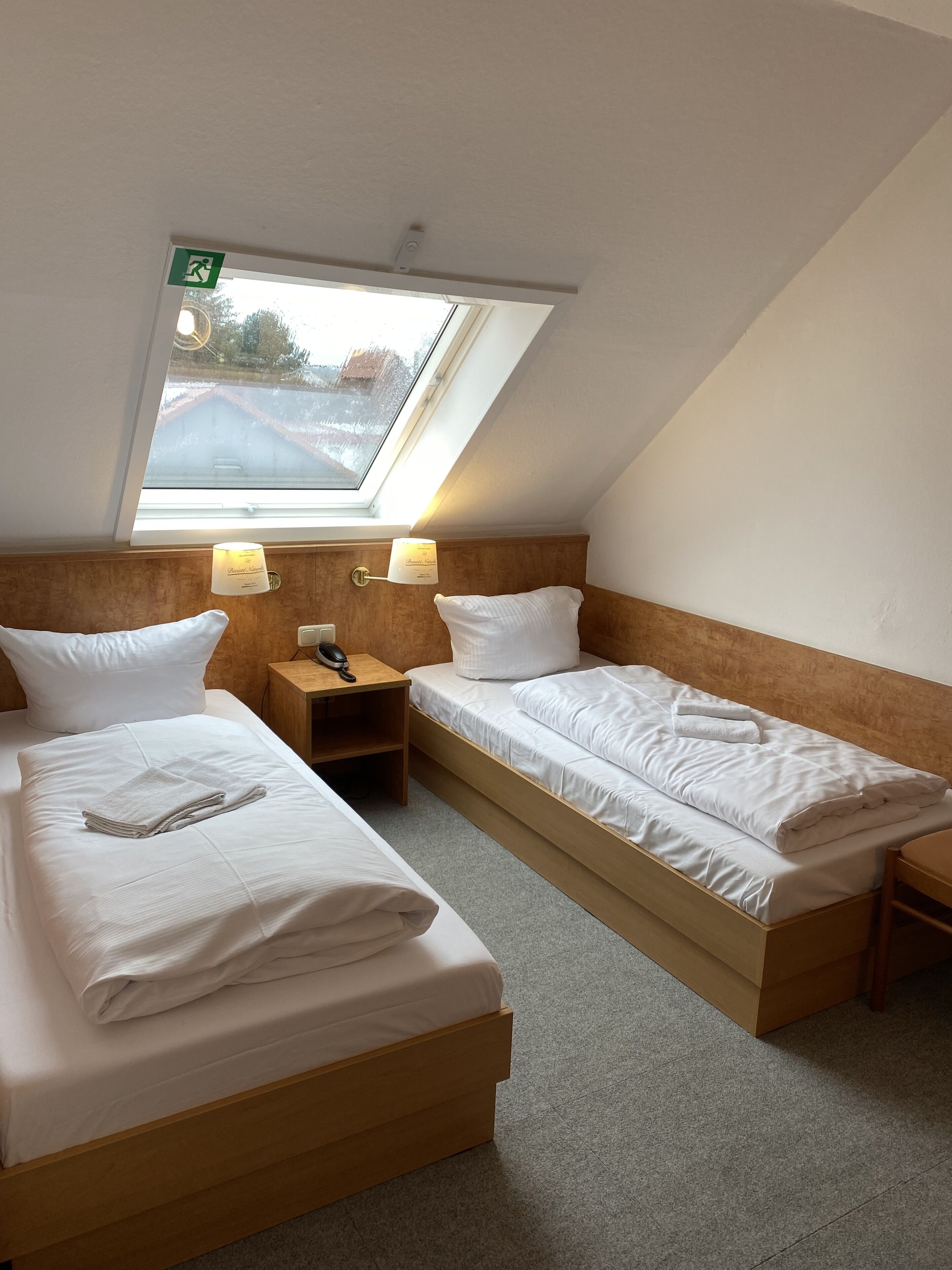 Une chambre avec deux lits simples, une petite table, une table de chevet, une lampe et une lucarne.