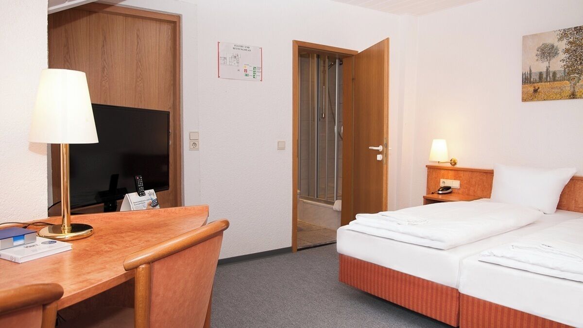 Una habitación de hotel con cama, escritorio, silla, lámpara y zona de ducha.