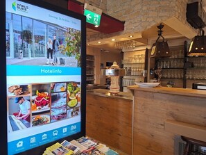 A hotel reception area with a digital screen displaying hotel information, a wooden reception desk, and a bar area with glasses and bottles. - Hotel Sonne (Karlsruhe)