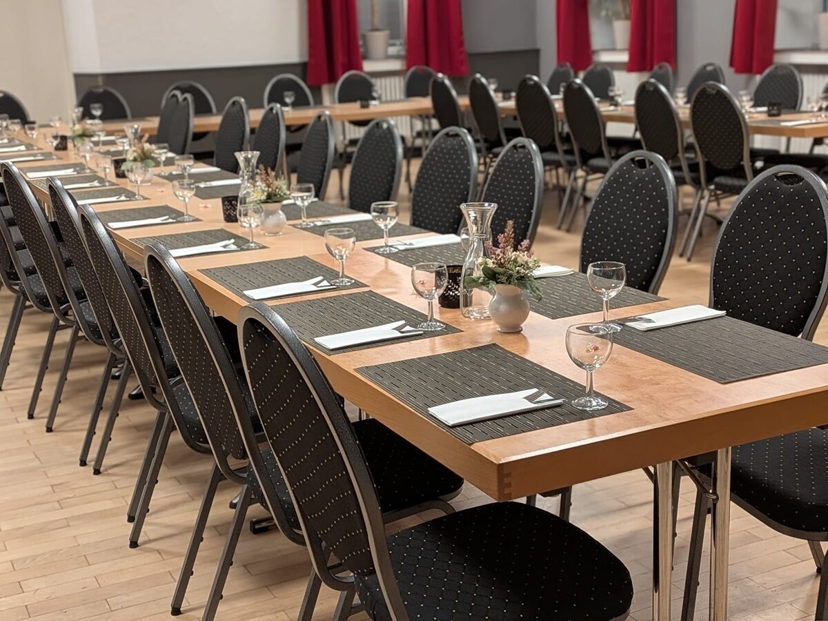 Sala de conferência com mesas longas, cadeiras pretas e louças postas.