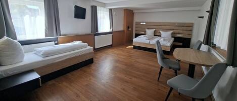 Habitación de hotel con suelo de madera, dos camas, una mesita redonda y un televisor colgado en la pared.