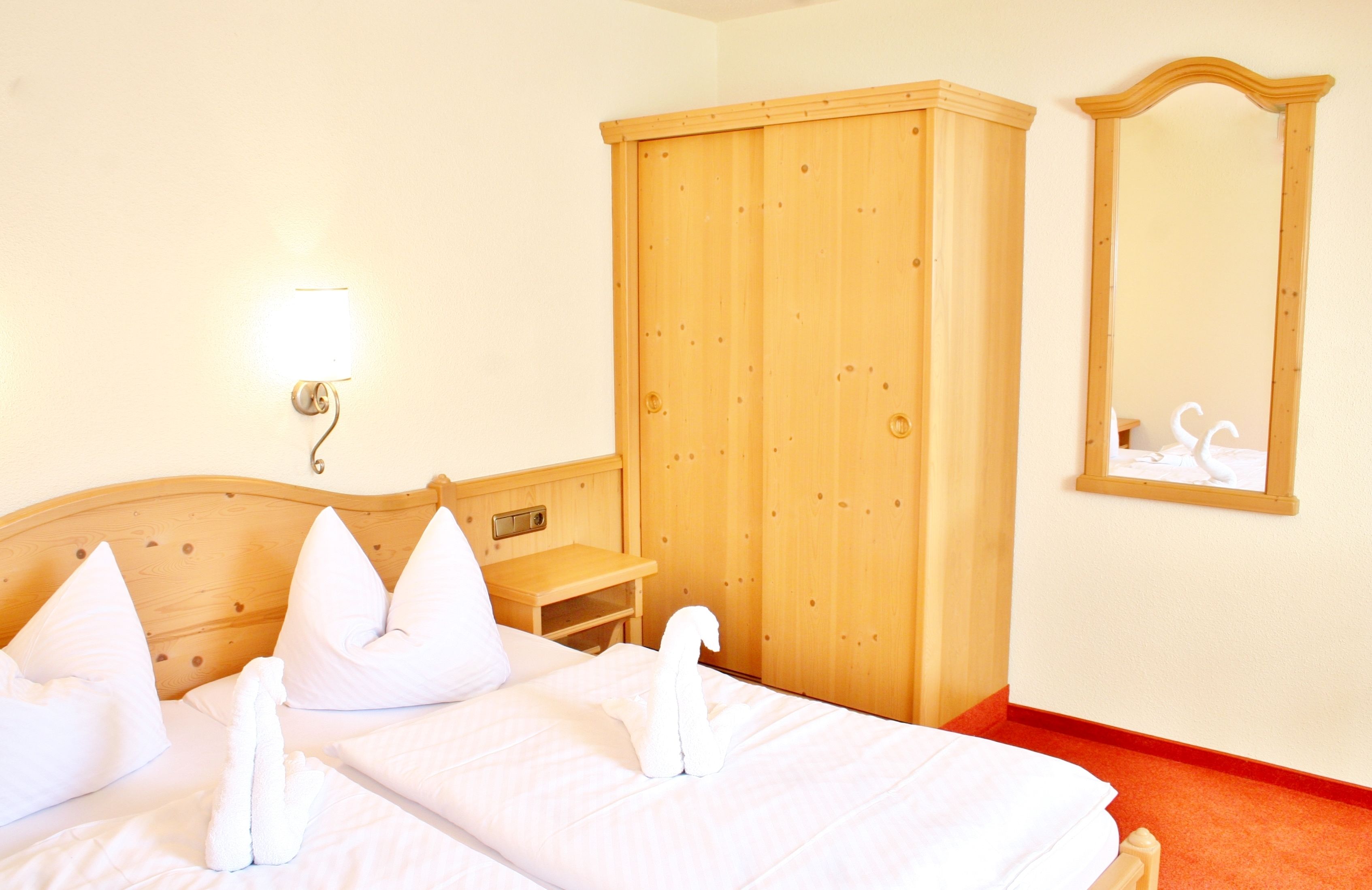 A hotel room with a wooden bed, white pillows, a wooden wardrobe, a bedside table, and a wall-mounted mirror.