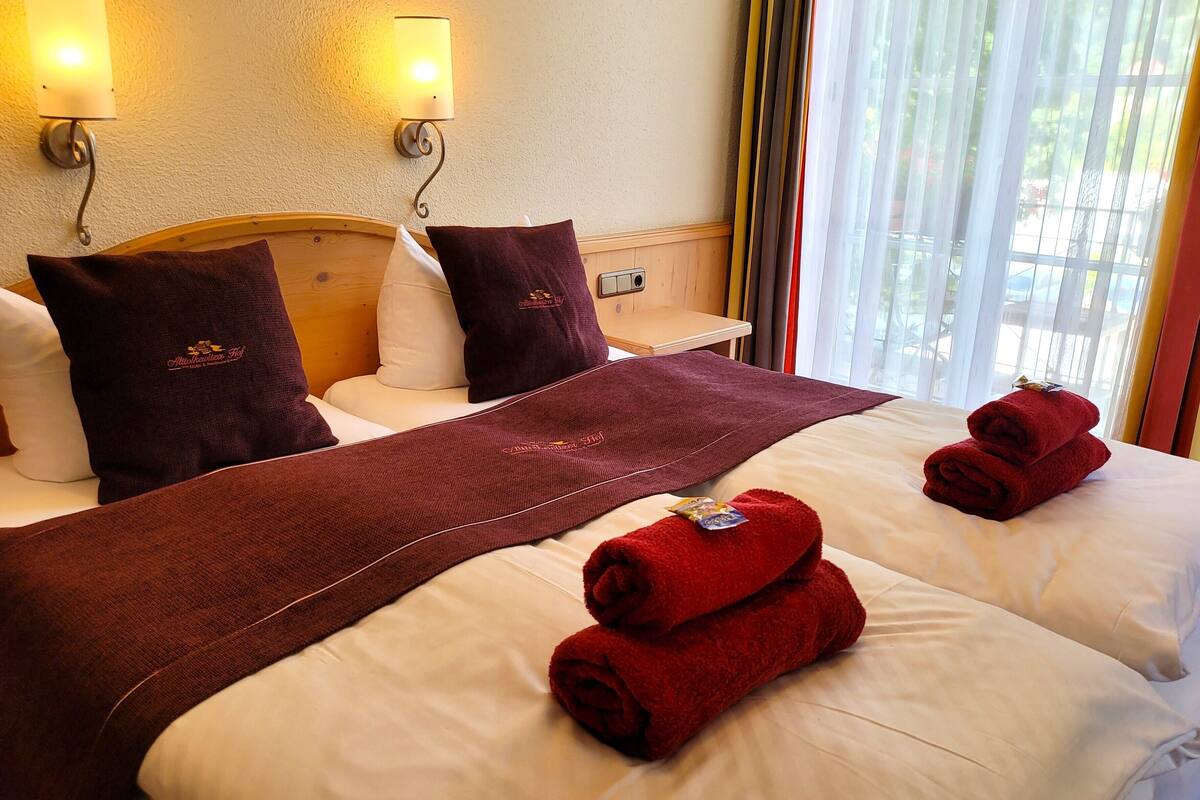 Habitación de hotel con dos camas, cada una con sábanas blancas y una manta roja. Dos lámparas de pared a cada lado de las camas, y una ventana con cortinas.