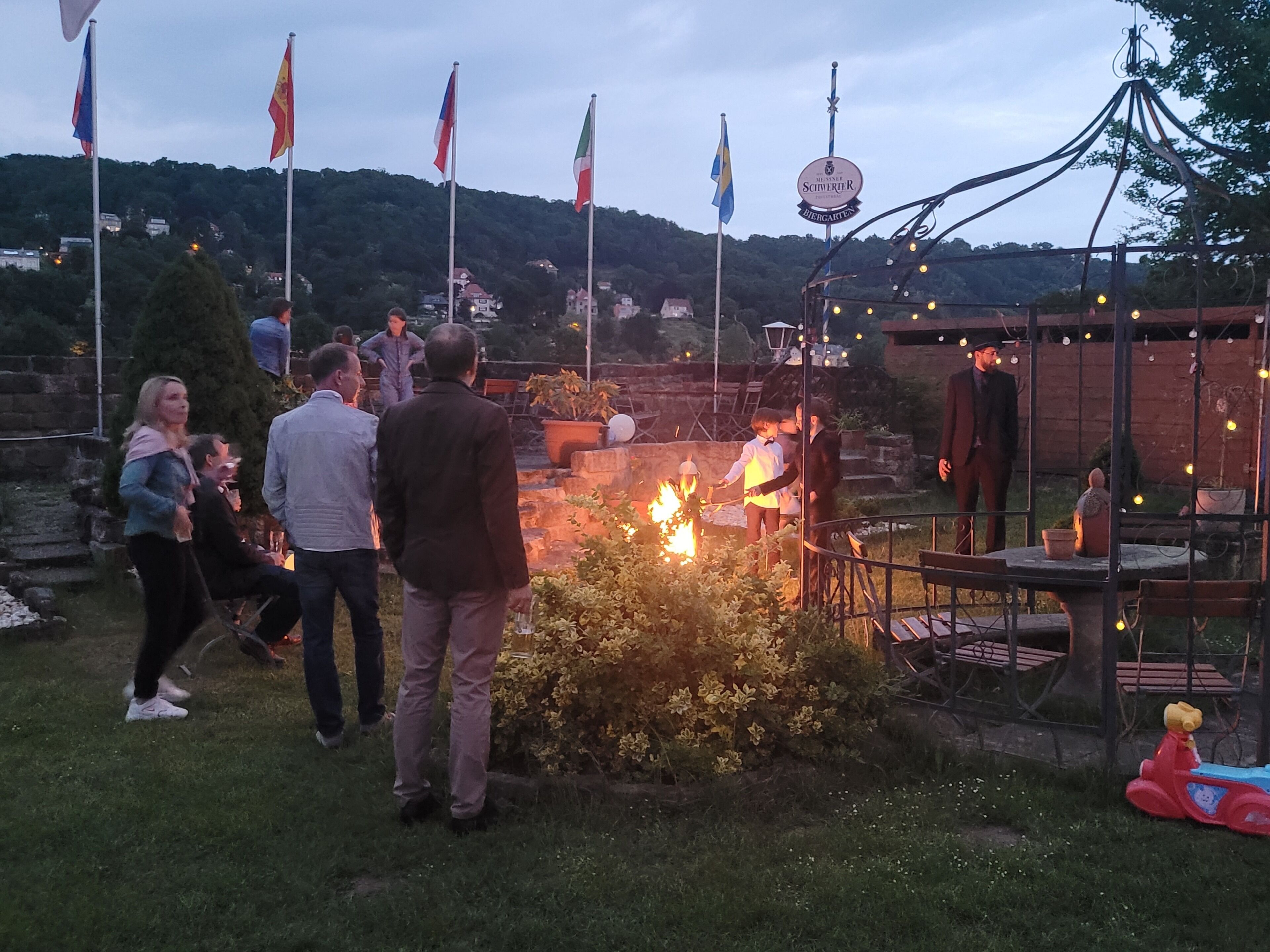 Eine Gruppe von Menschen hat sich im Freien auf einem Gelände mit mehreren Flaggen, einem Lagerfeuer und einem Schild für einen Biergarten versammelt.