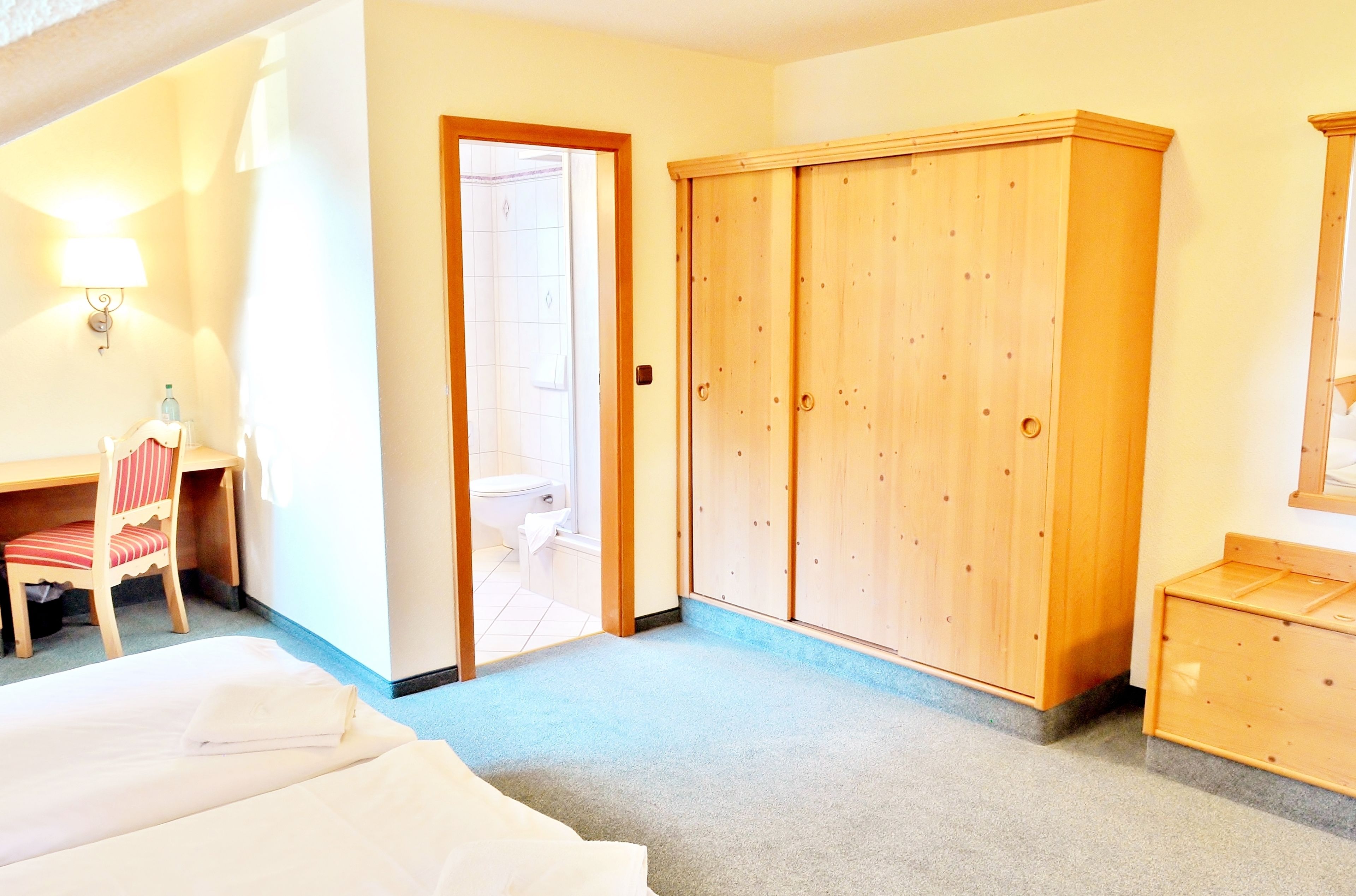 Ein Hotelzimmer mit einem Bett, einem Holzschrank, einem Schreibtisch mit Stuhl und einem durch eine offene Tür einsehbaren Badezimmer.