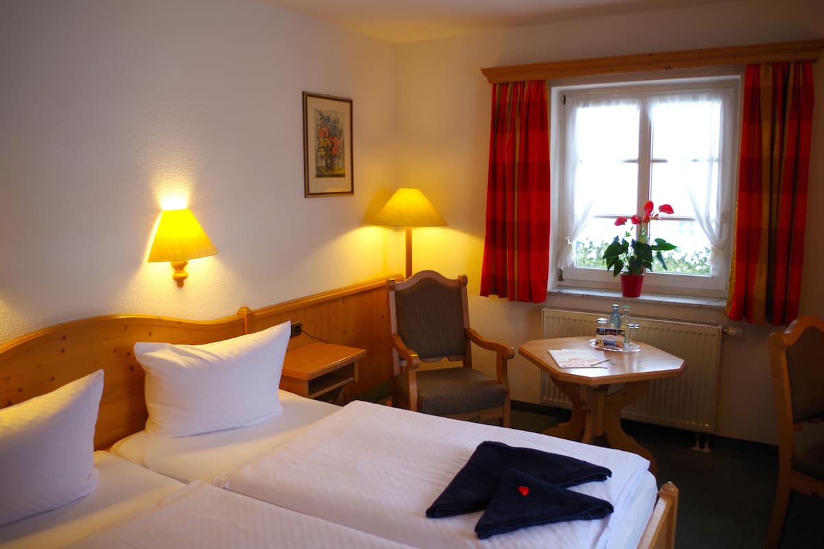 Una habitación de hotel con una cama, dos lámparas de noche, una silla, una mesita con una planta y una ventana con cortinas.
