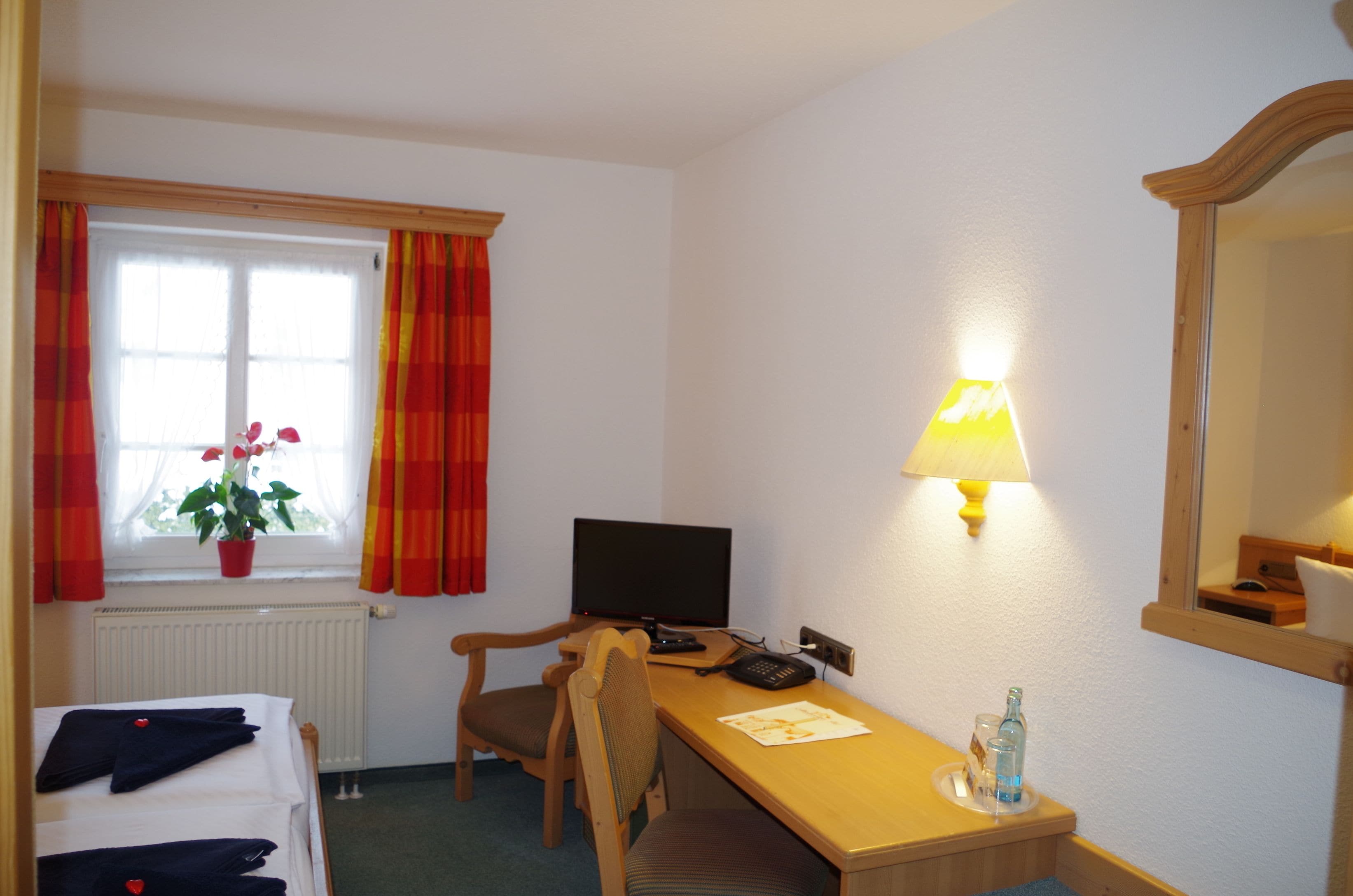 Een hotelkamer met een bed, een bureau met lamp, een stoel, een televisie, een plant en een raam met gordijnen.