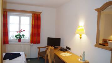 Een hotelkamer met een bed, een bureau met lamp, een stoel, een televisie, een plant en een raam met gordijnen.