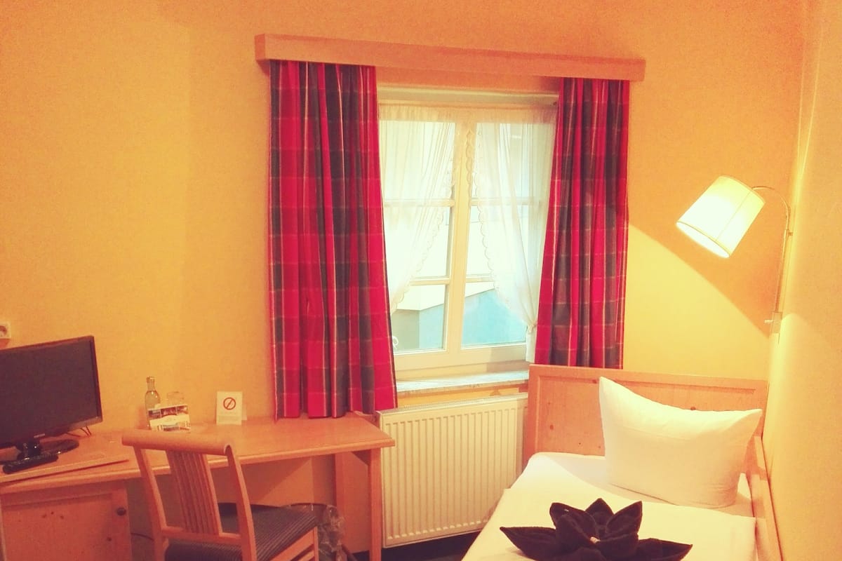 Una habitación de hotel con una cama individual, un escritorio con silla, televisión y ventana con cortinas.