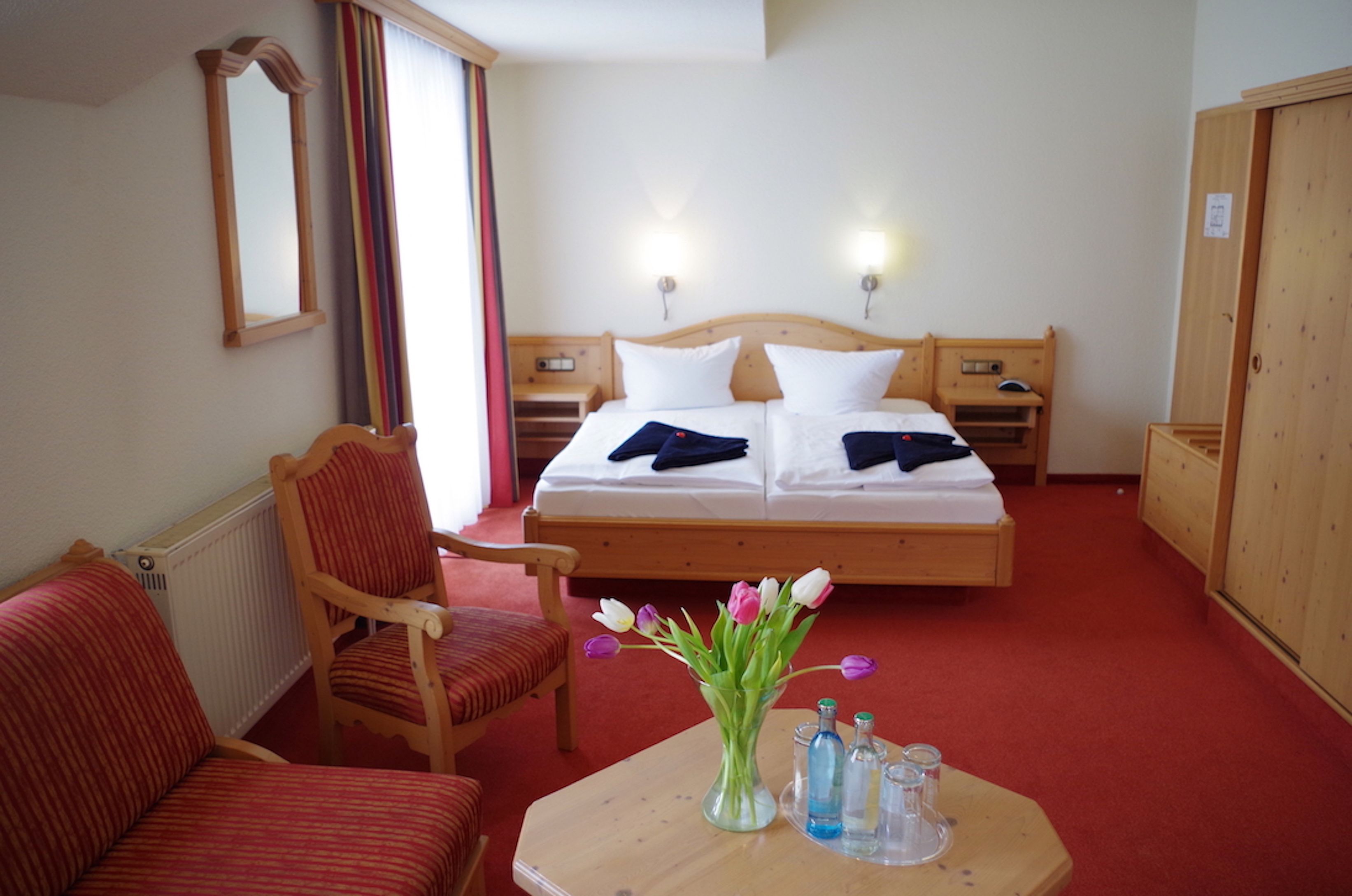 A hotel room with a bed, two bedside tables, a chair, a table with a vase and bottles, and a wardrobe.