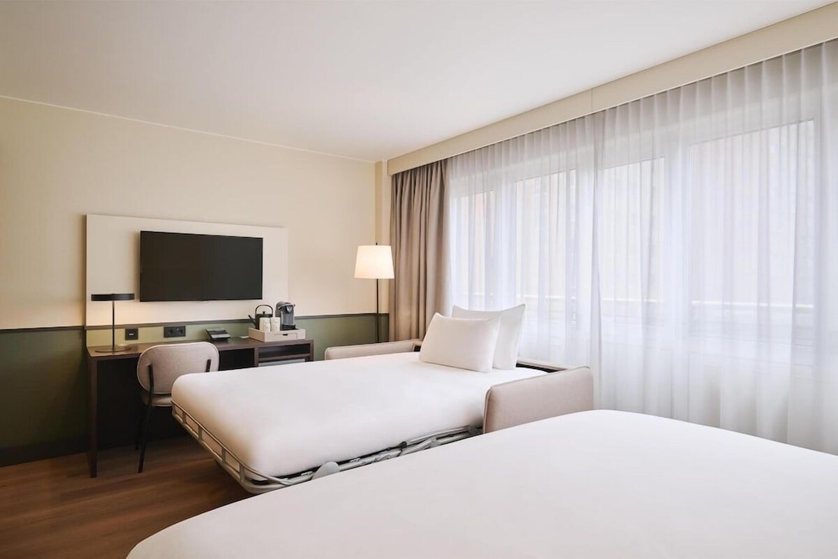 Quarto de hotel com duas camas, uma escrivaninha, uma cadeira, uma televisão e uma janela grande com cortinas.