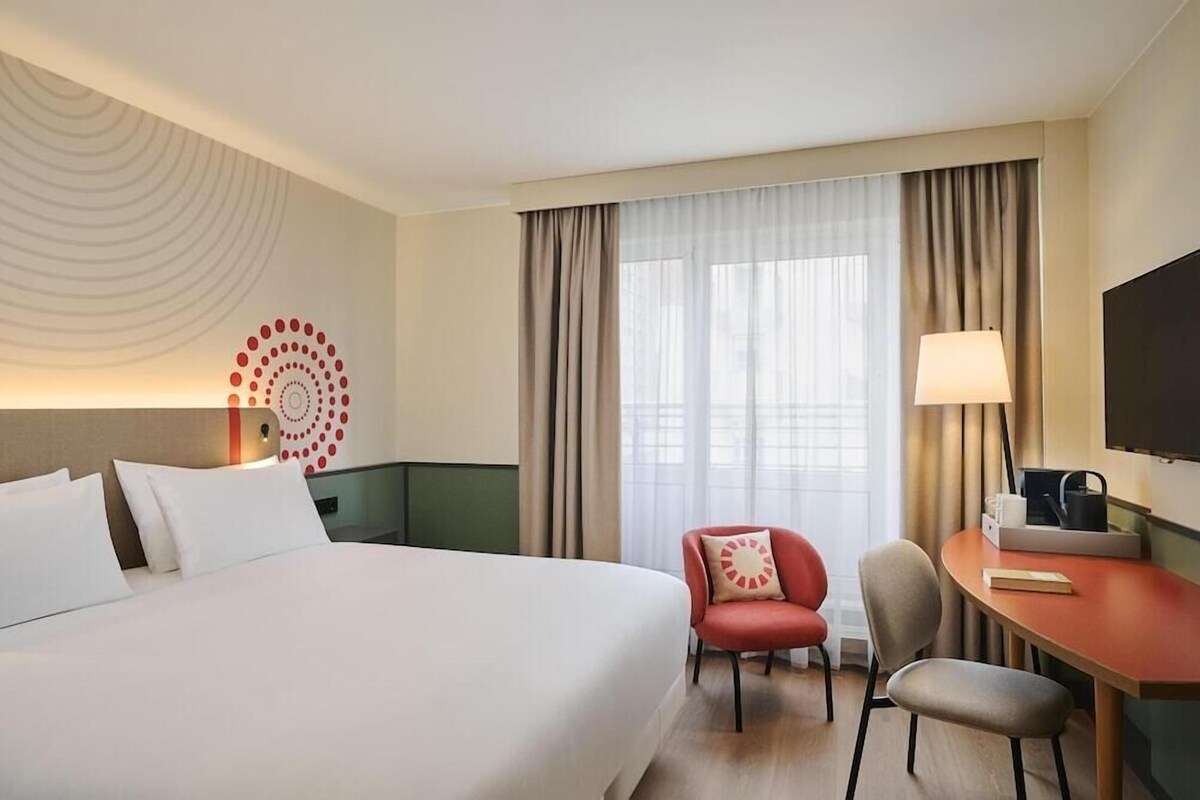 Une chambre d’hôtel avec un grand lit, un bureau avec une lampe, une télévision et une fenêtre avec des rideaux.