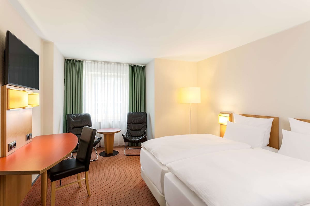 Une chambre d’hôtel avec un lit, un bureau, deux chaises, une télévision et une fenêtre avec des rideaux.