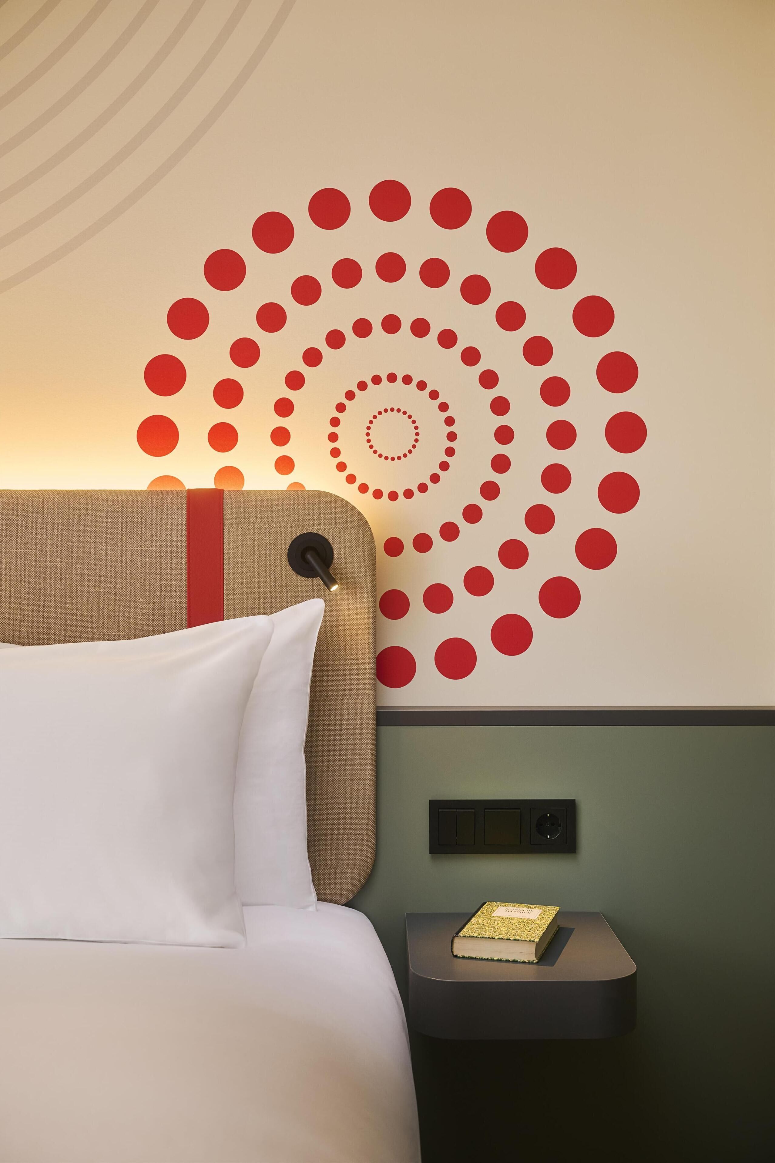 A hotel room with a bed, a wall with a circular pattern, a bedside table with a book, and a wall-mounted control panel.