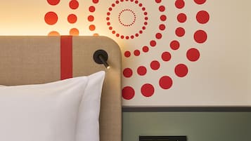 A hotel room with a bed, a wall with a circular pattern, a bedside table with a book, and a wall-mounted control panel.