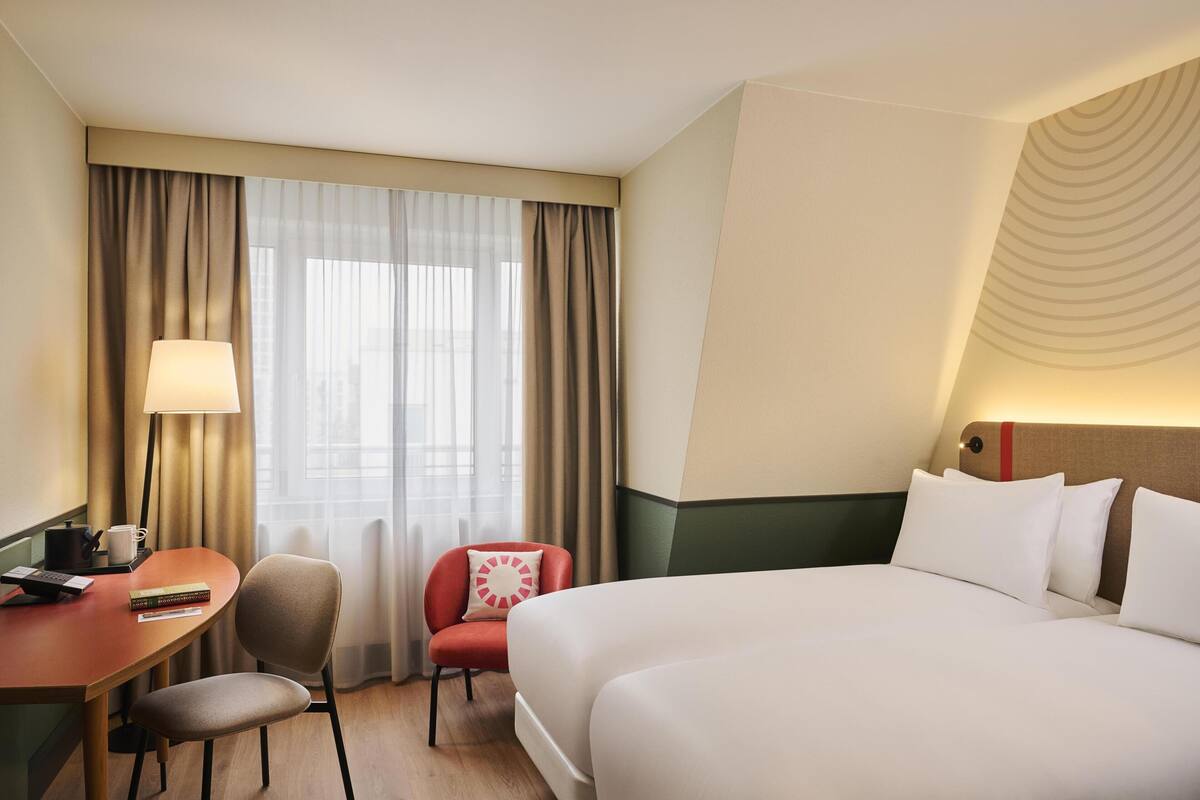 Une chambre d’hôtel avec un lit, un bureau, une chaise, une lampe et une fenêtre avec des rideaux.