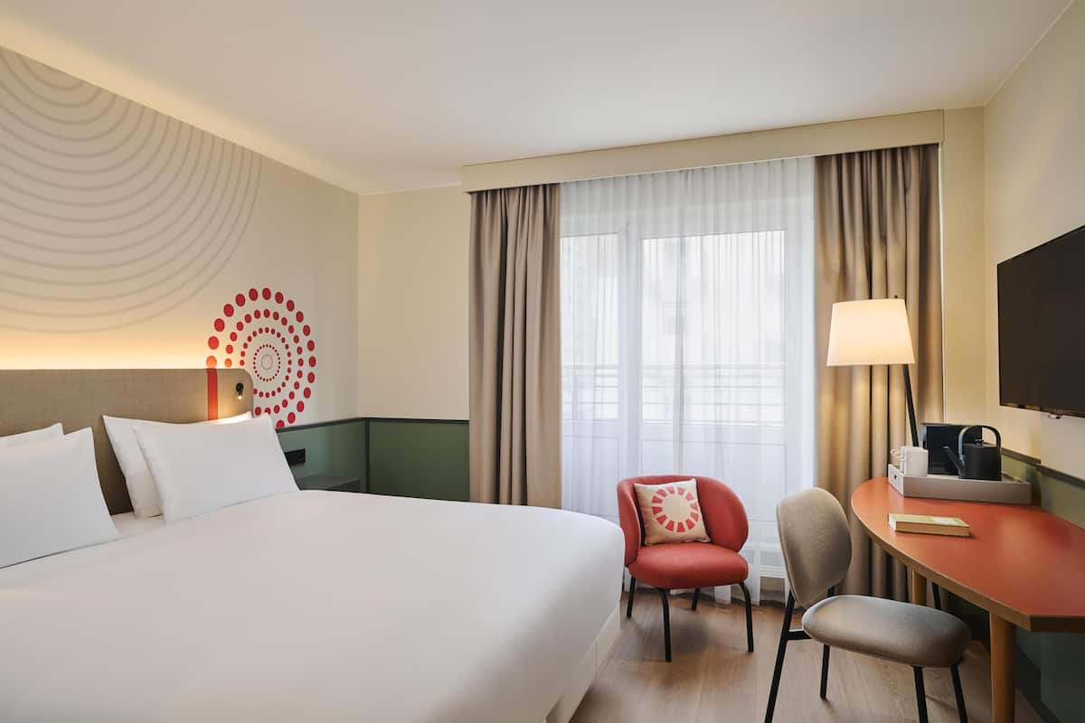 Une chambre d’hôtel équipée d’un lit, d’un bureau, d’une chaise, d’une télévision et d’une grande fenêtre avec des rideaux.