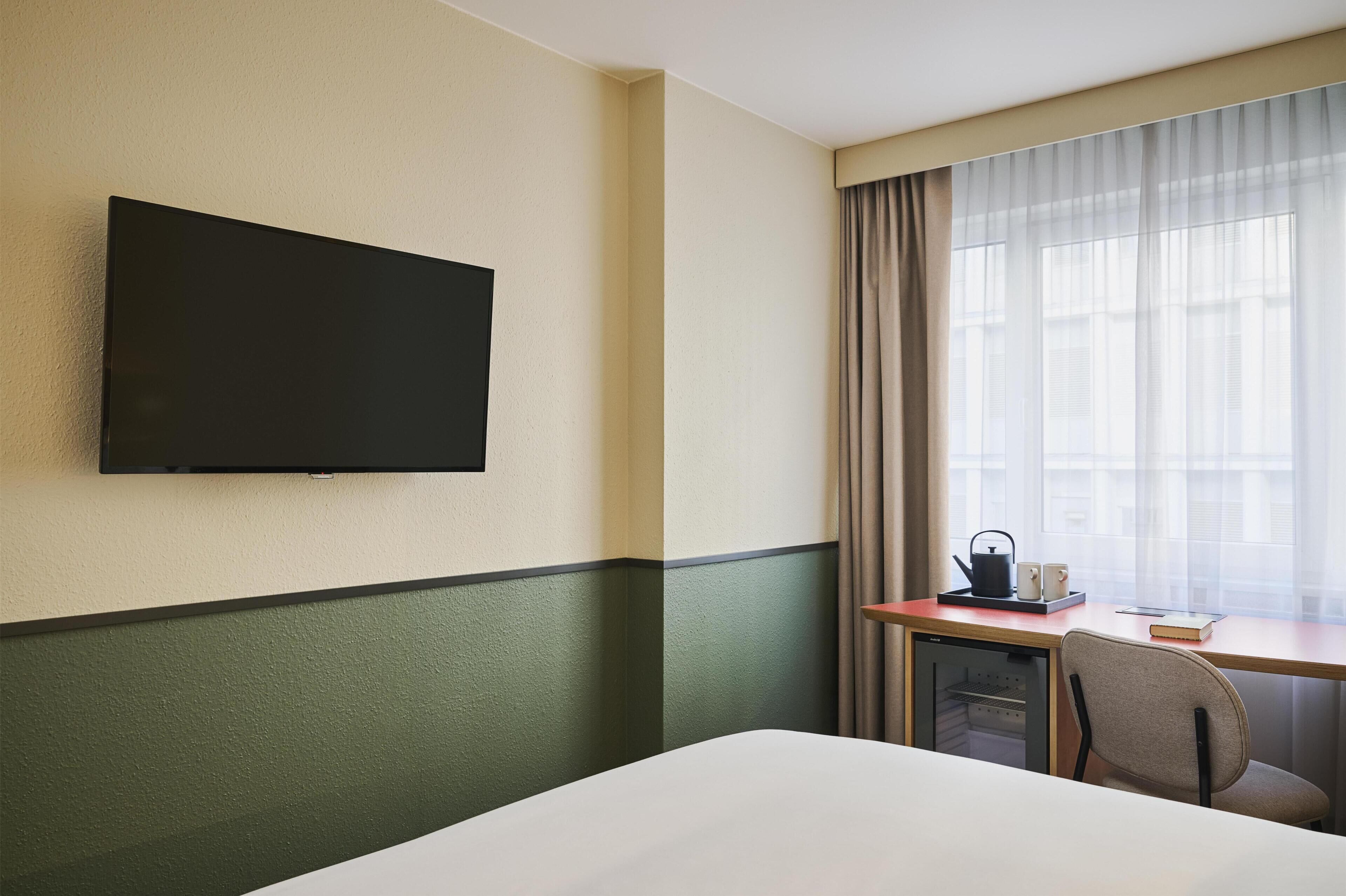 Una habitación de hotel con cama, escritorio y silla, televisor montado en la pared y ventana con cortinas.