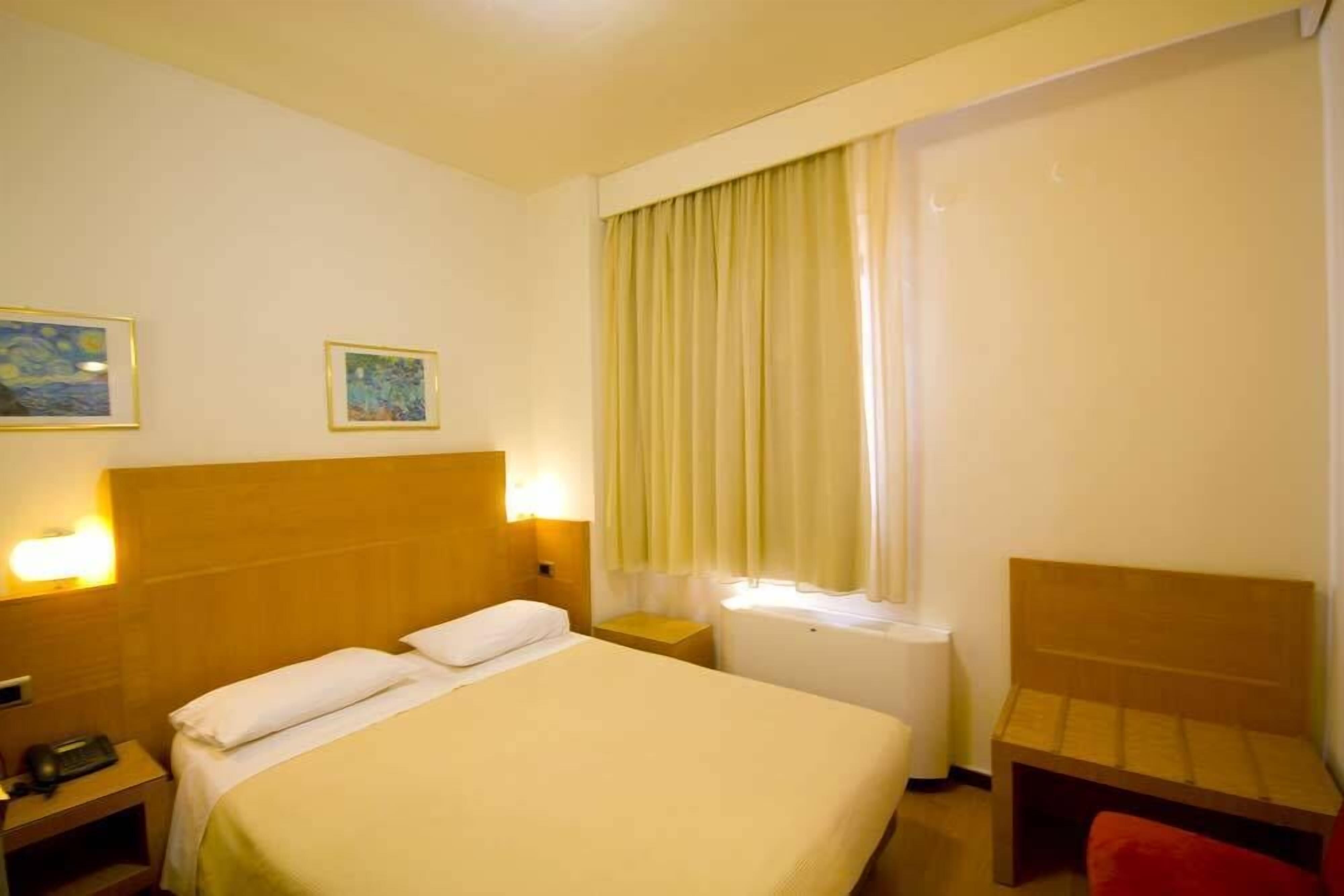 A hotel room with a bed, bedside tables, a nightstand, a desk, and a chair.