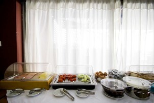 Montaje de bufé con diversos alimentos en recipientes transparentes, cubiertos sobre platos y una ventana con cortinas traslúcidas al fondo.