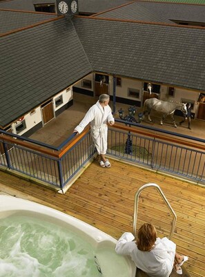 A man in a white robe standing on a wooden deck overlooking a stable with horses.