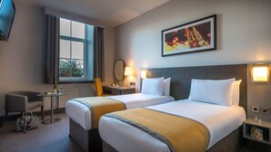 Habitación de hotel con dos camas, una ventana, un escritorio, una silla y un cuadro en la pared.