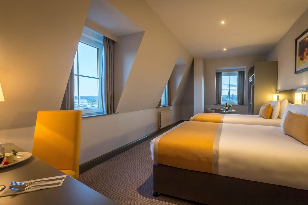 Een hotelkamer met twee bedden, een bureau met stoel, een eettafel met bestek en uitzicht op een stadsgezicht door de ramen.