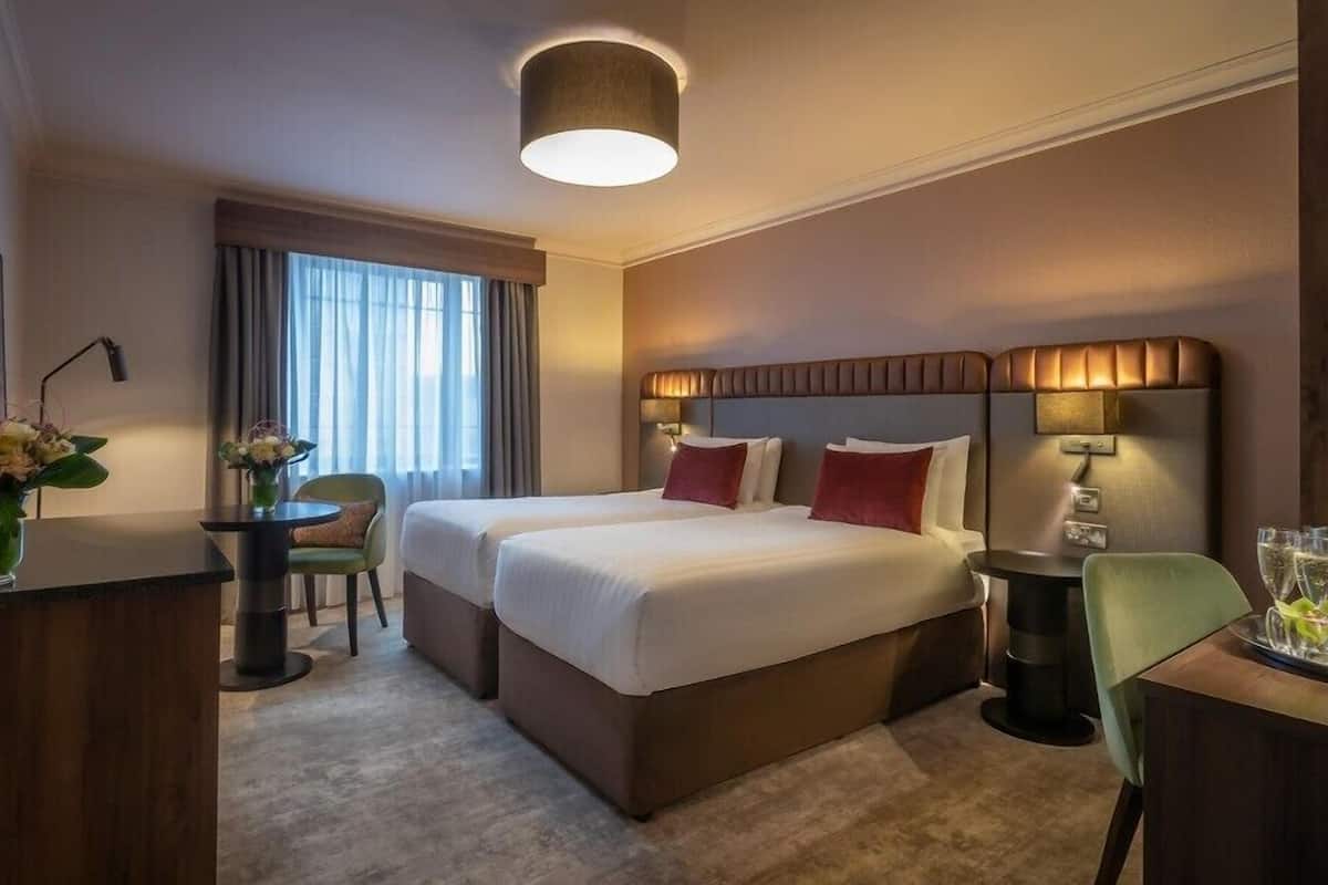 A hotel room with a double bed, a desk, a chair, and a small table with a vase of flowers.