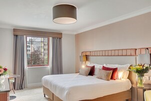 Una habitación de hotel con una cama grande, una ventana con cortinas, una mesa redonda con un florero y una lámpara en la mesita de noche.