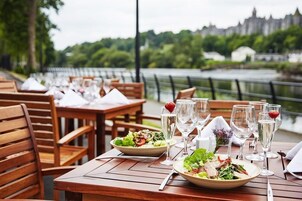 Un espacio al aire libre para comer con mesas y sillas de madera, frente a un río y vegetación.