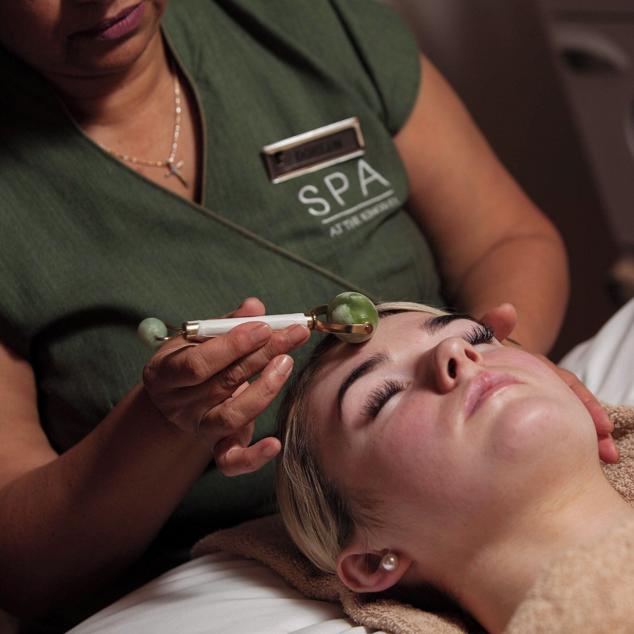 Una persona recibe un tratamiento facial con un instrumento, en una habitación con una cama y una pared verde.