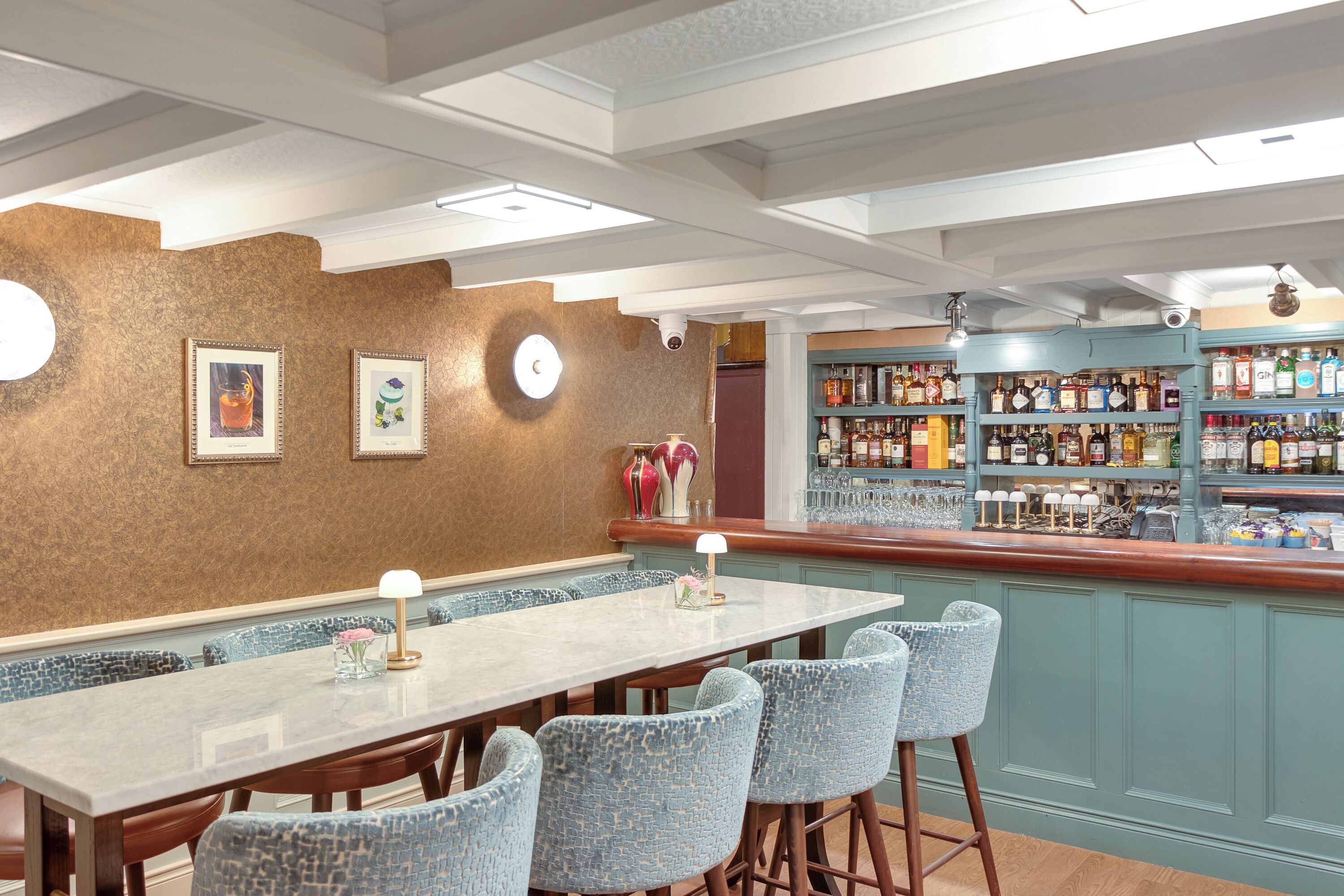 A bar area with a long table and blue chairs, a wall with framed pictures, and shelves with liquor bottles.