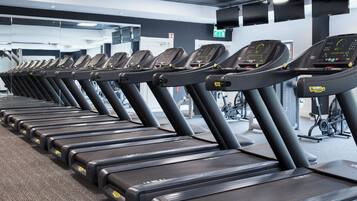 Varias caminadoras en un gimnasio con espejos en las paredes y equipo de ejercicio al fondo.
