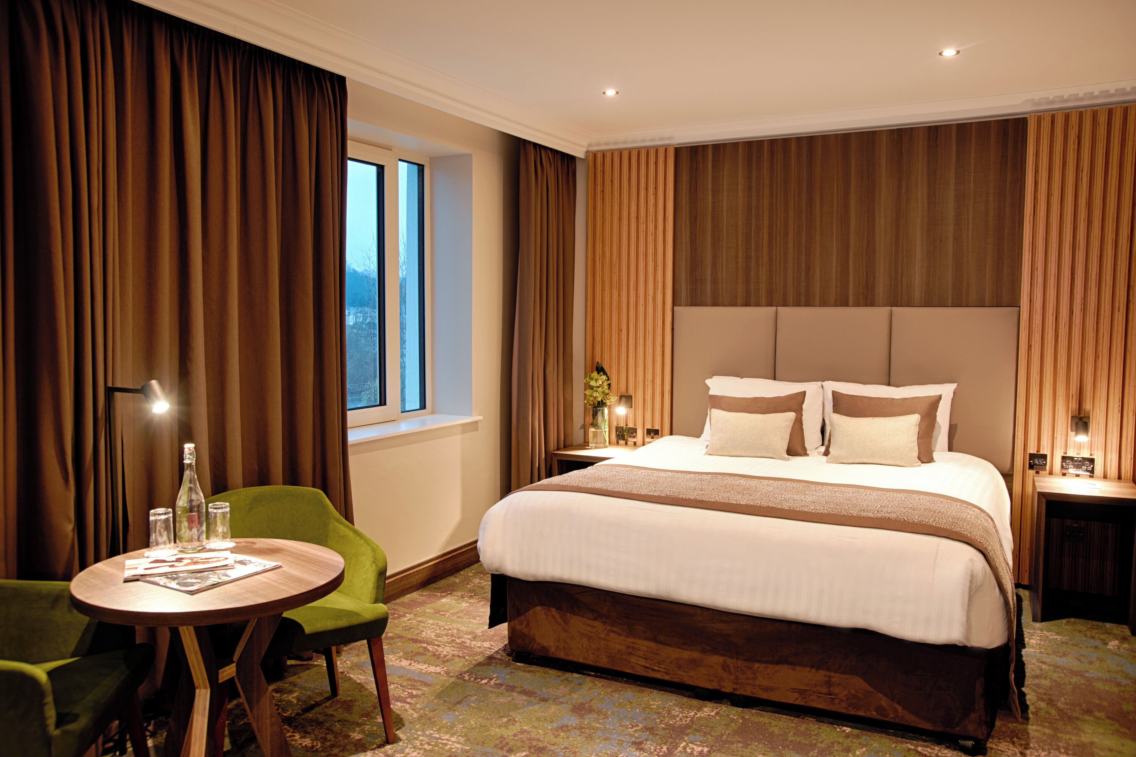 A hotel room with a large bed, a round wooden table, a green armchair, and a window with curtains.