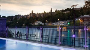 Ein Schwimmbecken mit Metallzaun, einer Laterne und Blick auf ein historisches Gebäude im Hintergrund.