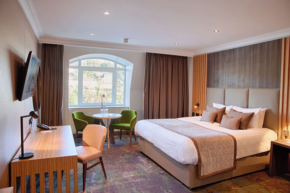 A hotel room with a large bed, a desk with a chair, a small round table with two chairs, and a TV mounted on the wall.