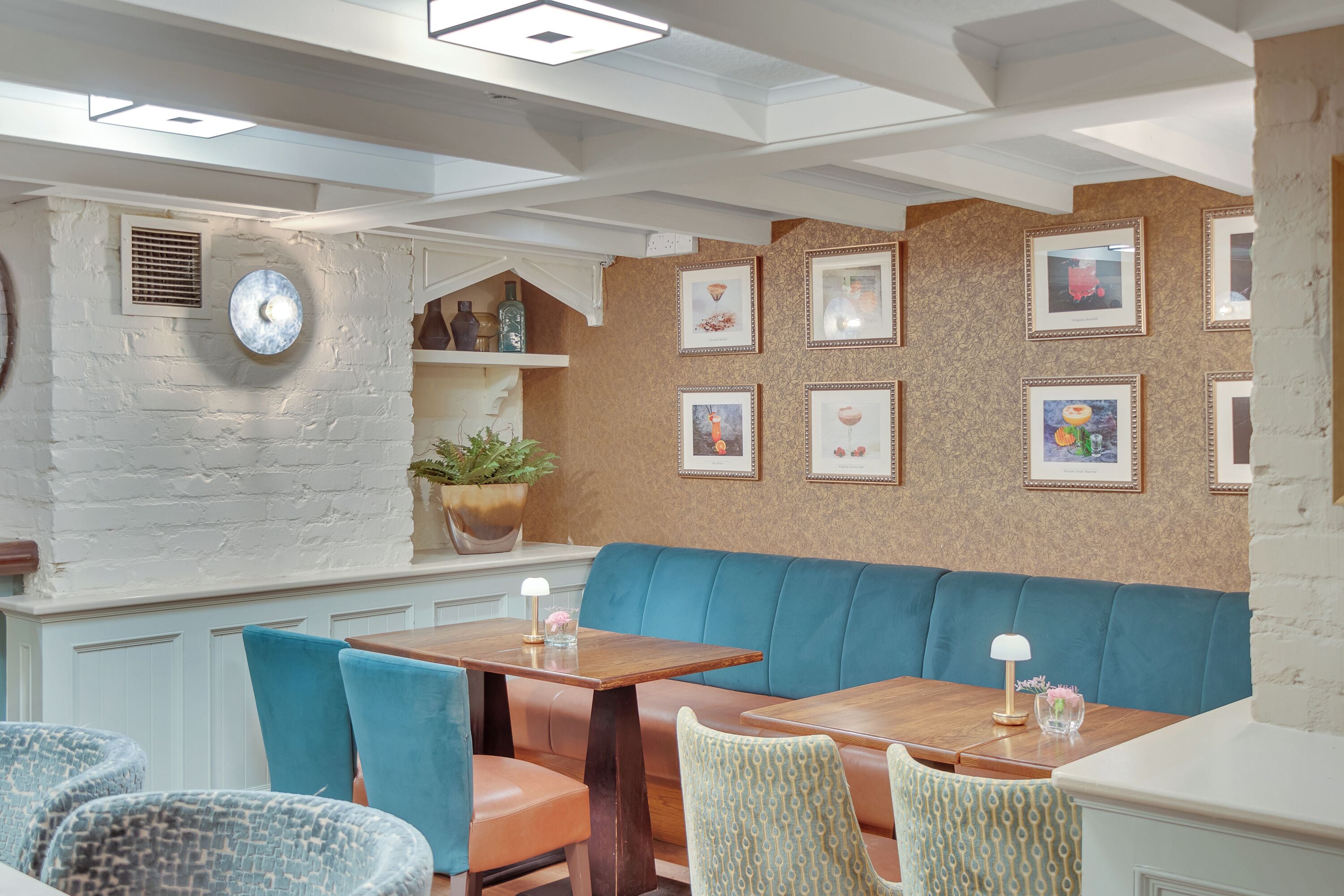 A restaurant with a blue cushioned bench, wooden tables, and chairs. There are framed pictures on the wall and a potted plant on a shelf.