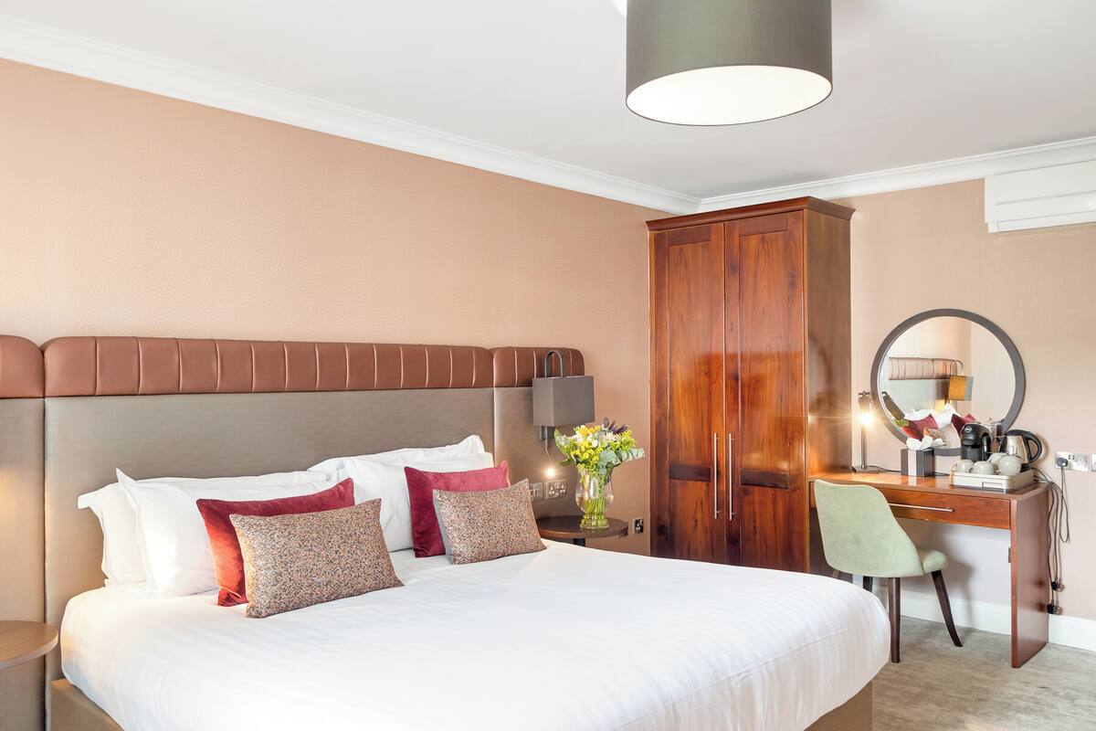 A hotel room with a large bed, a wooden wardrobe, a desk with a chair, and a mirror.