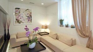 Een moderne woonkamer met een beige bank, een bijzettafeltje met een bloevas, een schilderij aan de muur en een raam met gordijnen.