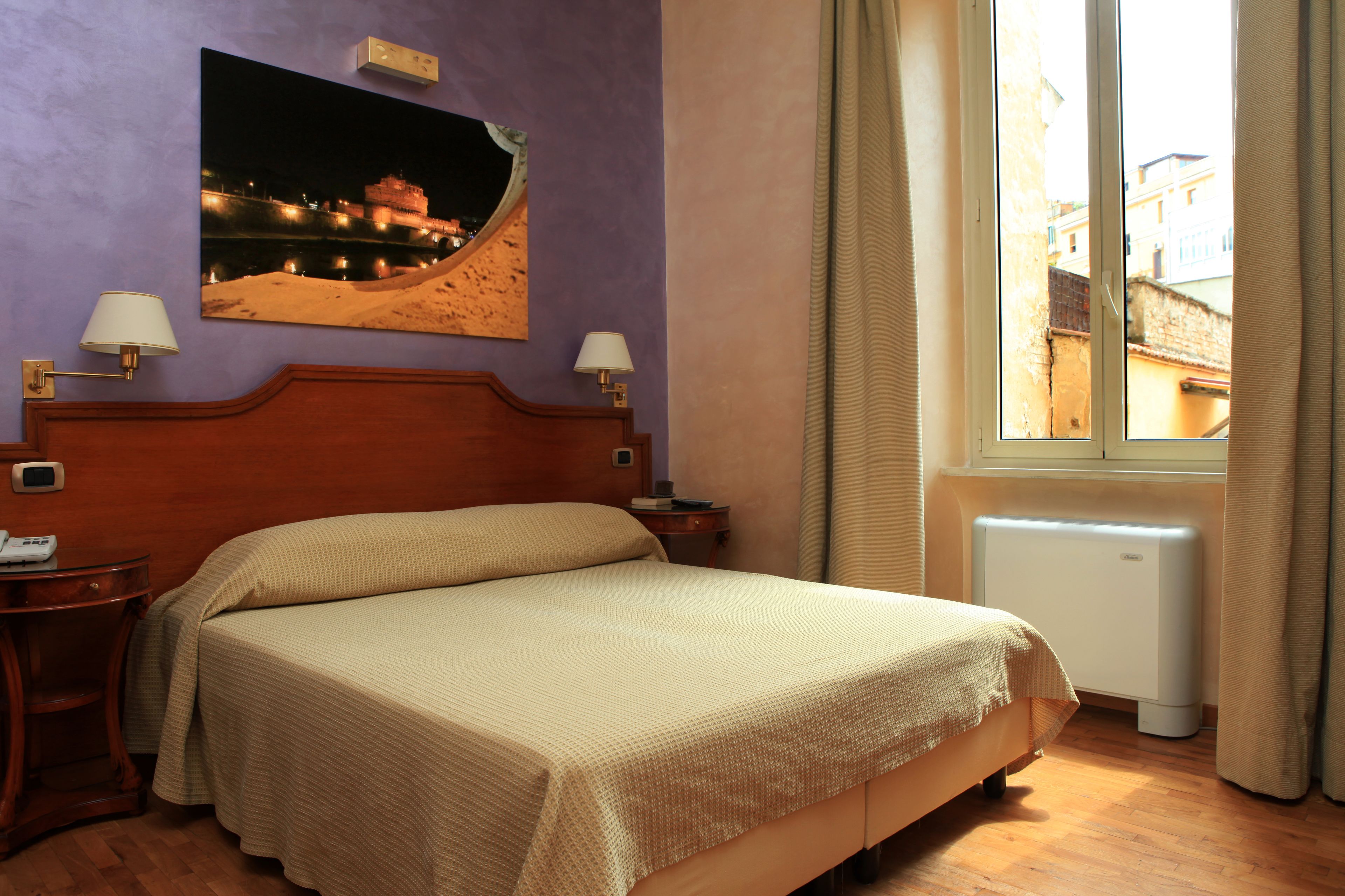 A hotel room with a bed, two bedside lamps, a nightstand, a wall-mounted picture, and a window with curtains.