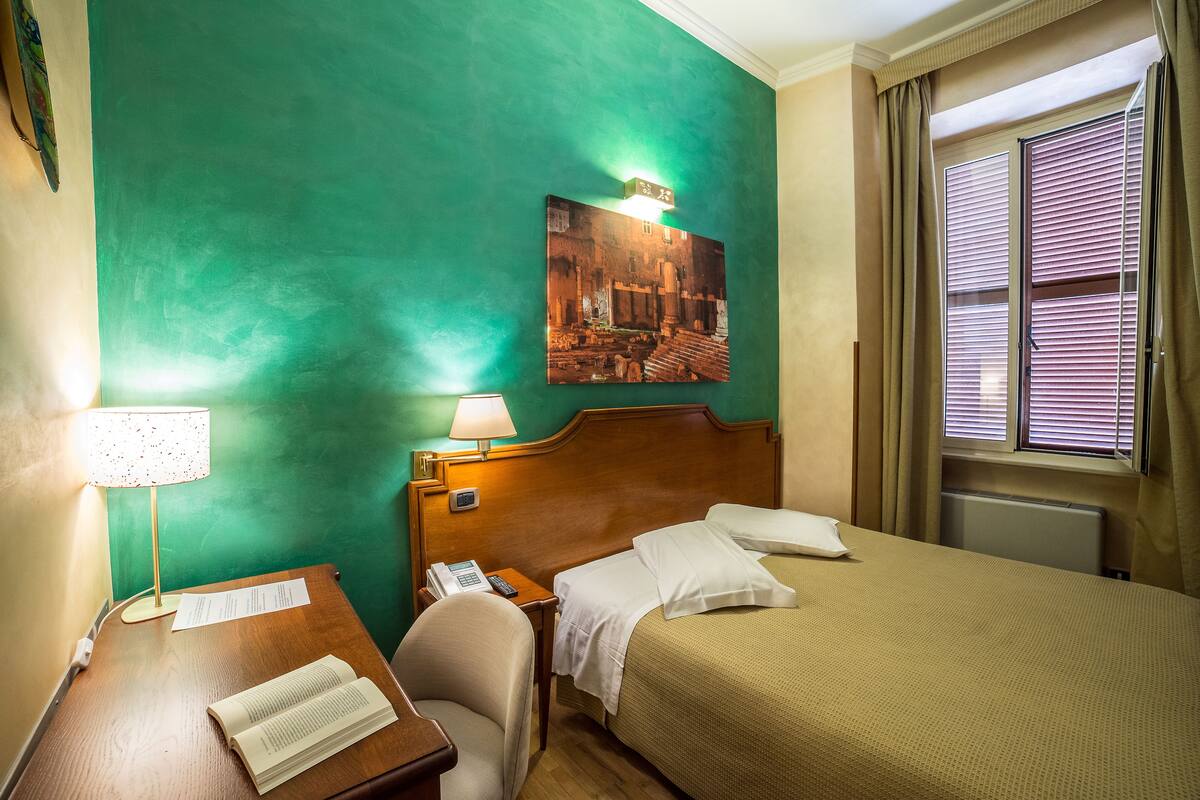 Habitación de hotel con cama, escritorio con lámpara, silla, un cuadro en la pared y ventana con persianas.
