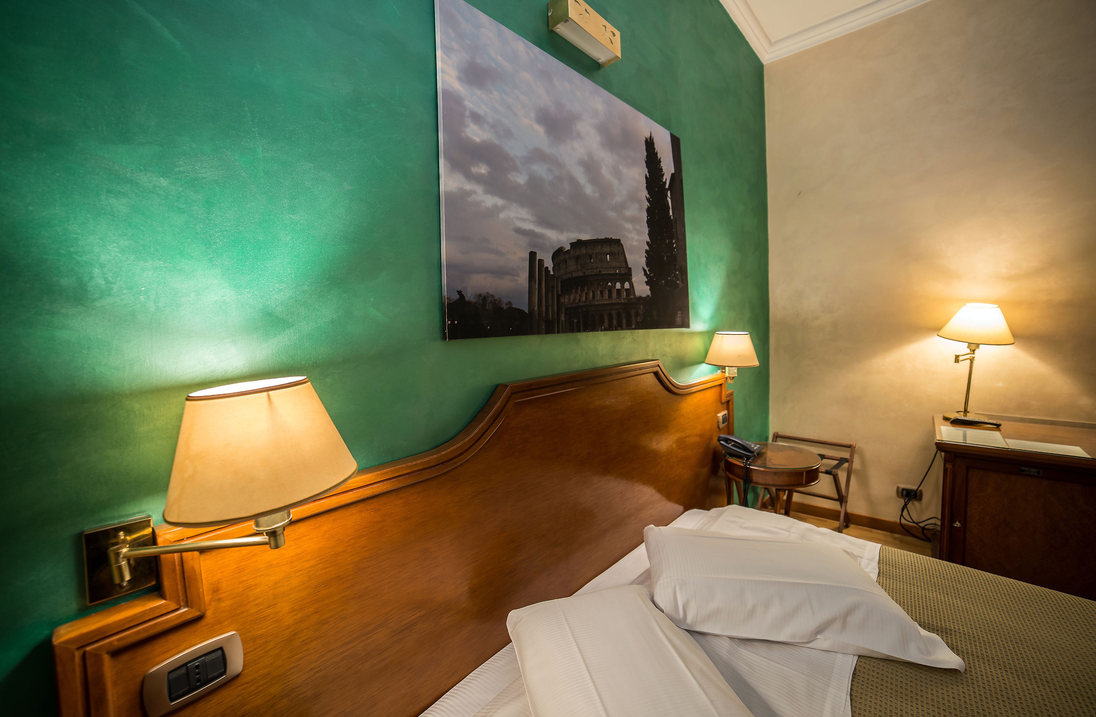 Ein Hotelzimmer mit einem Holzkopfteil, zwei Nachttischlampen, einem Gemälde eines historischen Gebäudes, einem kleinen Tisch mit einem Stuhl und einem Schreibtisch mit Lampe.