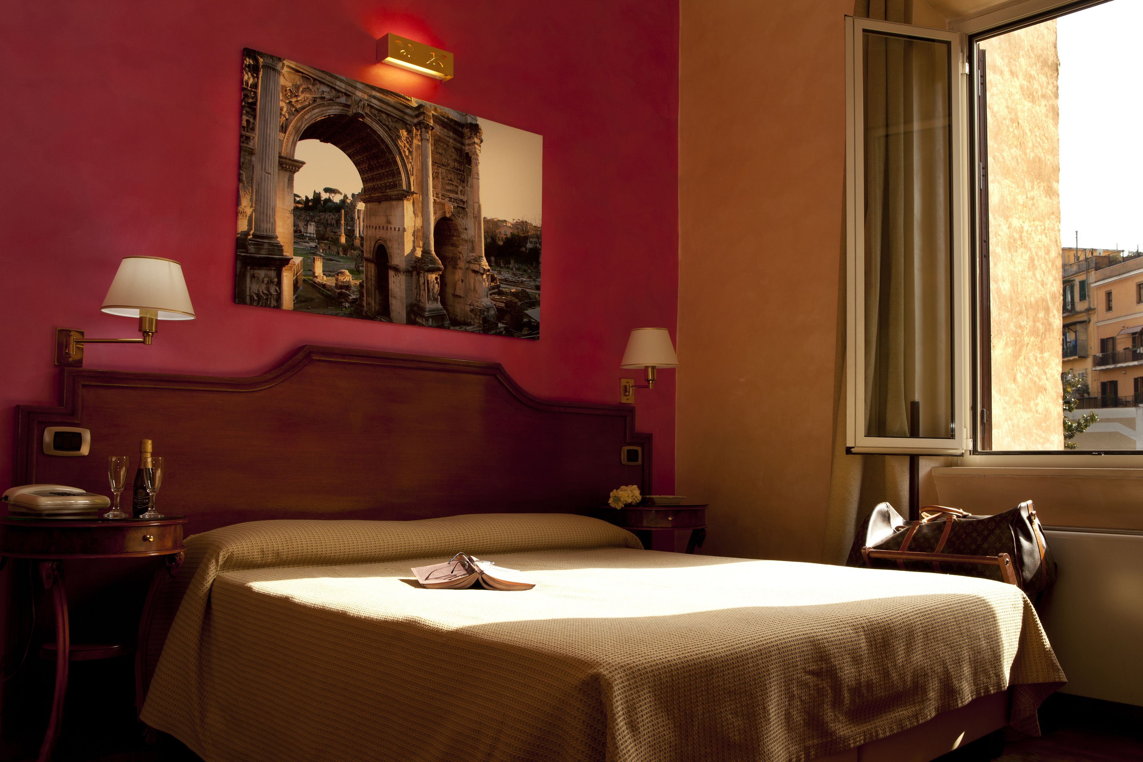 A hotel room with a large bed, two bedside tables, a nightstand, a window with a view, and a wall-mounted picture of an ancient archway.