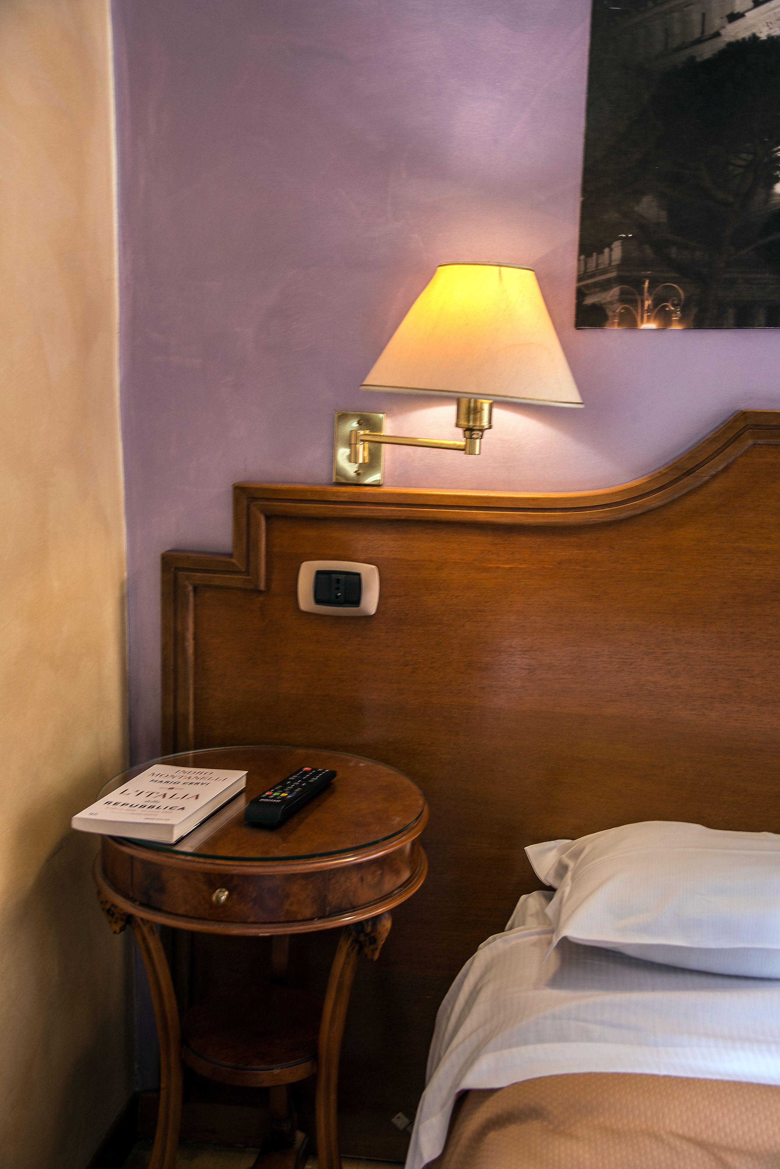 A hotel room with a wooden headboard, a bedside table with a remote control and a book, and a wall-mounted lamp.