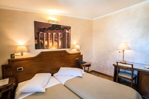 Habitació d'hotel amb un llit doble, dues taules de nit amb làmpades, un escriptori petit amb una cadira i un quadre penjat a la paret amb arquitectura antiga.
