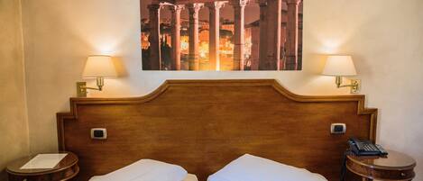 Hotelkamer met een houten hoofdeinde, twee nachtkastjes met lampen, een bed met witte lakens, en een lijst met daarop een afbeelding van oude kolommen.