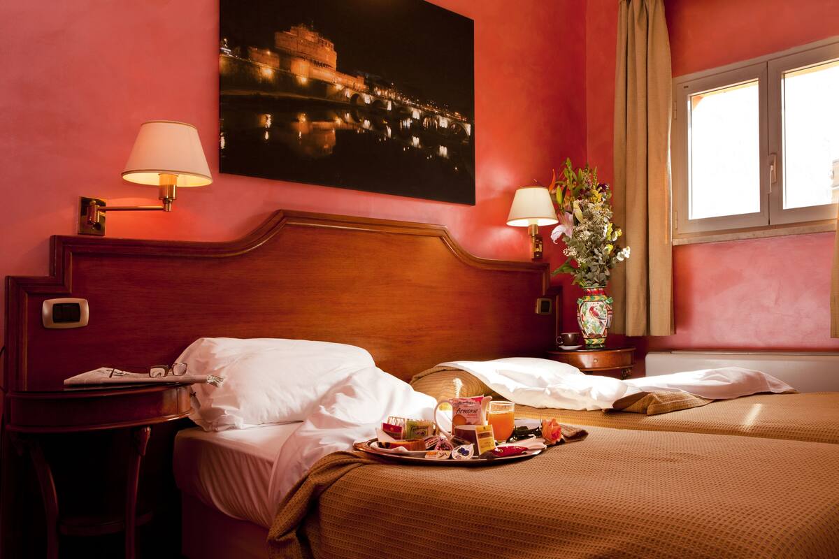 Habitación de hotel con dos camas, una bandeja de desayuno, una pintura con vista nocturna y un jarrón con flores.