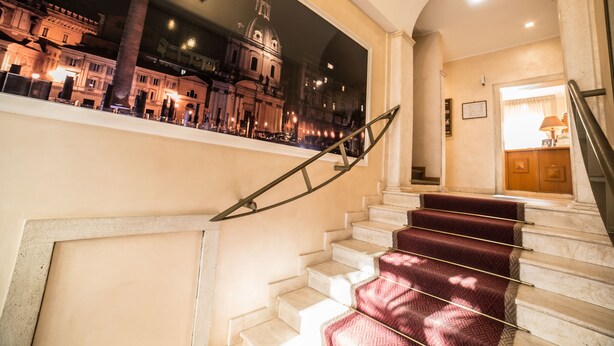 Una imponente escalera de mármol con alfombra roja, que conduce a un espacio bien iluminado con un amplio ventanal con vistas a un edificio histórico.