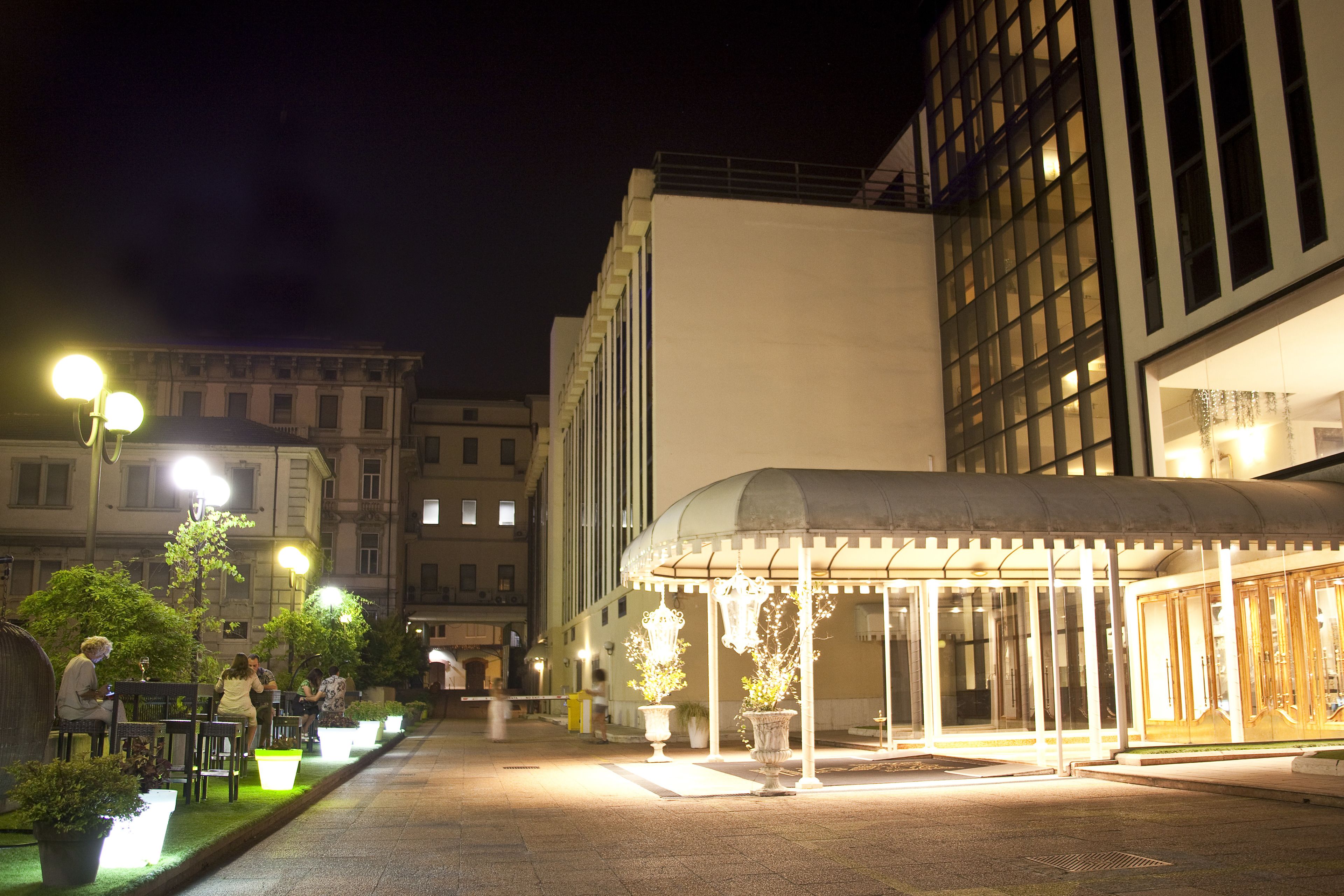 A nighttime urban square with a covered seating area, potted plants, and street lamps.